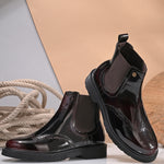 A glossy burgundy Chelsea boot with brogue detailing, elastic side panels, and a durable black sole, featuring a gold accent logo.