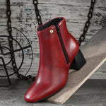 Suede ankle boot with pointed toe, chunky block heel, side zipper, and gold logo detail near the top.