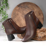 Suede ankle boot with pointed toe, chunky block heel, side zipper, and gold logo detail near the top.