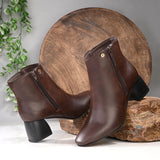 Suede ankle boot with pointed toe, chunky block heel, side zipper, and gold logo detail near the top.