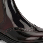 A glossy Wine Chelsea boot with brogue detailing, elastic side panels, and a durable black sole, featuring a gold accent logo.