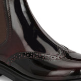 A glossy Wine Chelsea boot with brogue detailing, elastic side panels, and a durable black sole, featuring a gold accent logo.