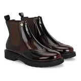 A glossy burgundy Chelsea boot with brogue detailing, elastic side panels, and a durable black sole, featuring a gold accent logo.