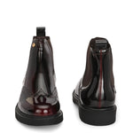 A glossy Wine Chelsea boot with brogue detailing, elastic side panels, and a durable black sole, featuring a gold accent logo.