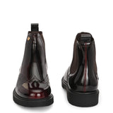 A glossy Wine Chelsea boot with brogue detailing, elastic side panels, and a durable black sole, featuring a gold accent logo.