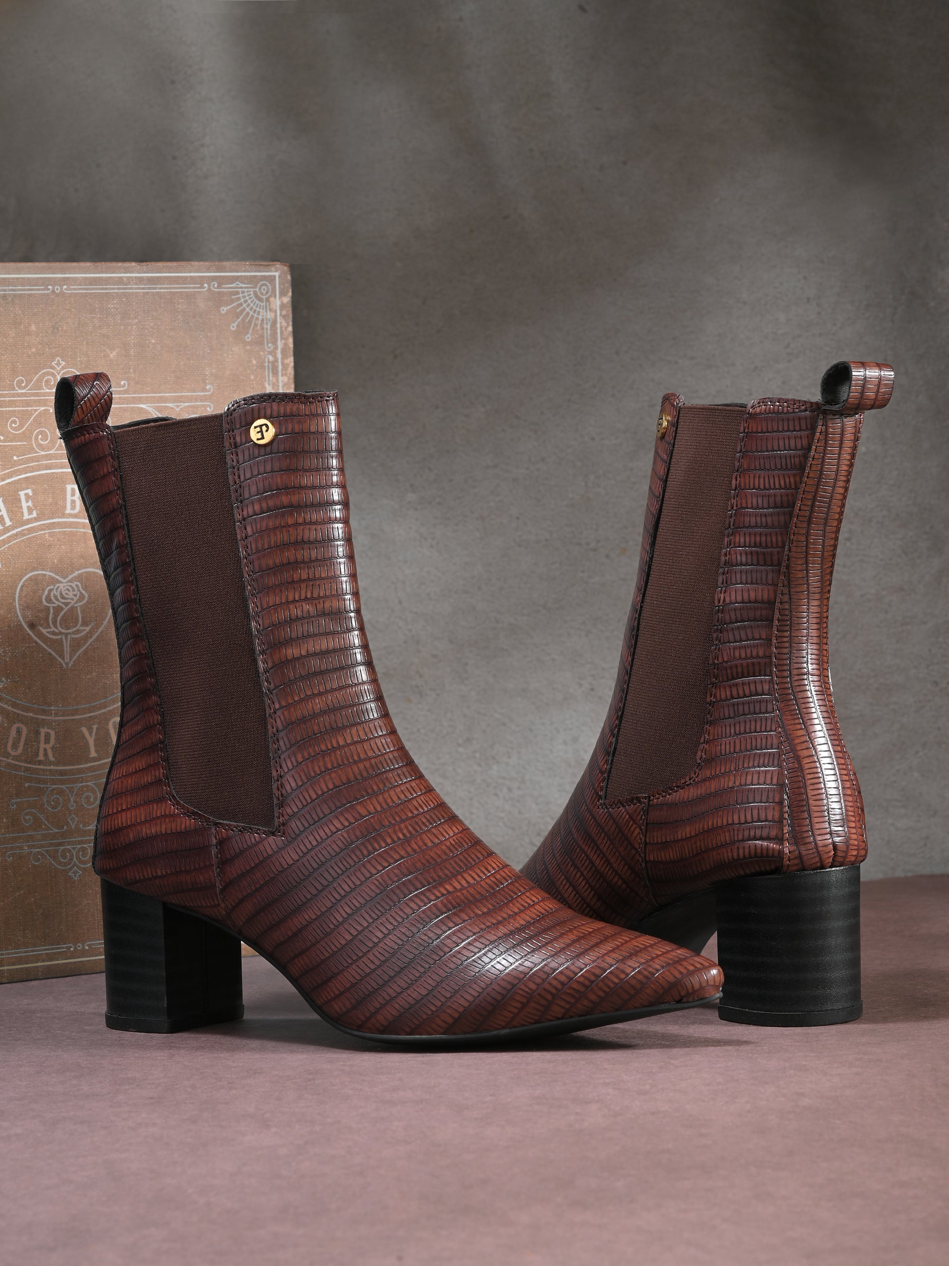 A sleek Tan crocodile-textured ankle boot with a pointed toe and mid-block heel, featuring an elastic side panel and a gold accent logo.