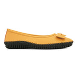 Mustard yellow leather flat shoe with a decorative bow, black textured sole, and visible stitching for a casual, stylish look.