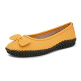 Mustard yellow leather flat shoe with a decorative bow, black textured sole, and visible stitching for a casual, stylish look.