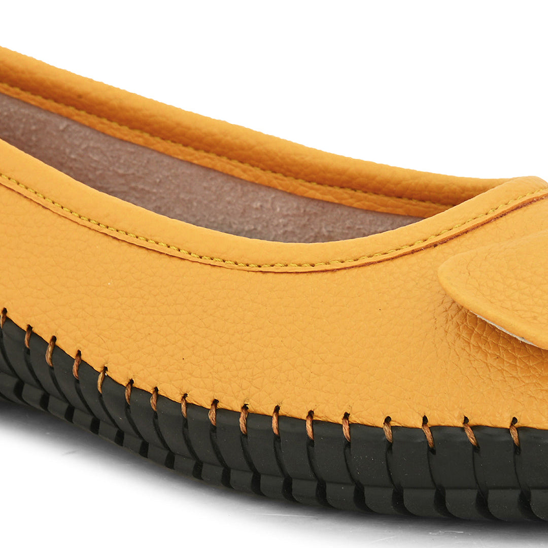 Mustard yellow leather flat shoe with a decorative bow, black textured sole, and visible stitching for a casual, stylish look.